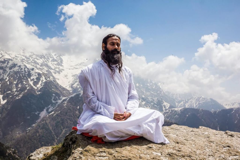 An image of a man in a meditative pose, seated on a mountain, surrounded by a serene natural landscape.